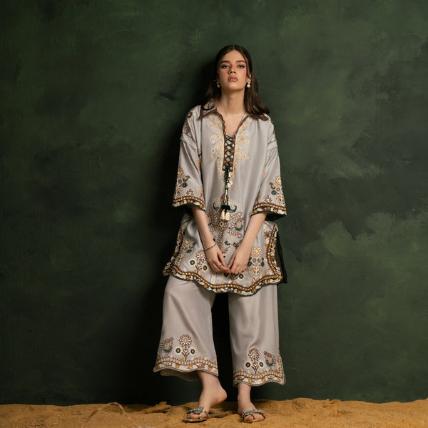 A woman models a light gray three-piece ethnic wear set featuring a long-sleeve embroidered tunic, wide-leg trousers, and a matching belt, set against a dark green textured backdrop with a sandy floor and woven baskets.