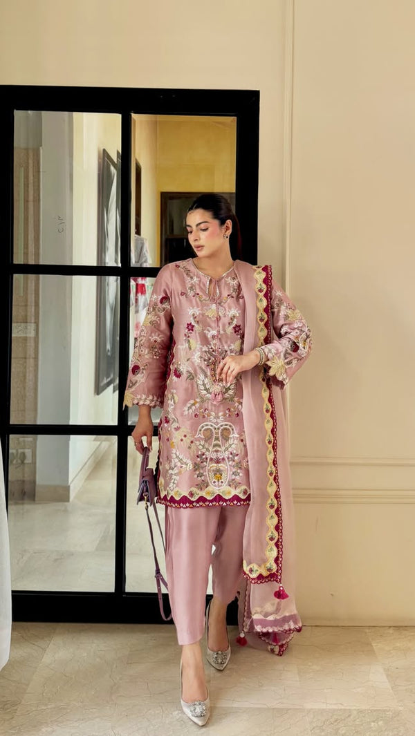 A woman models a pink embroidered traditional outfit with matching pants and dupatta in front of a beige wall and black-framed glass door.