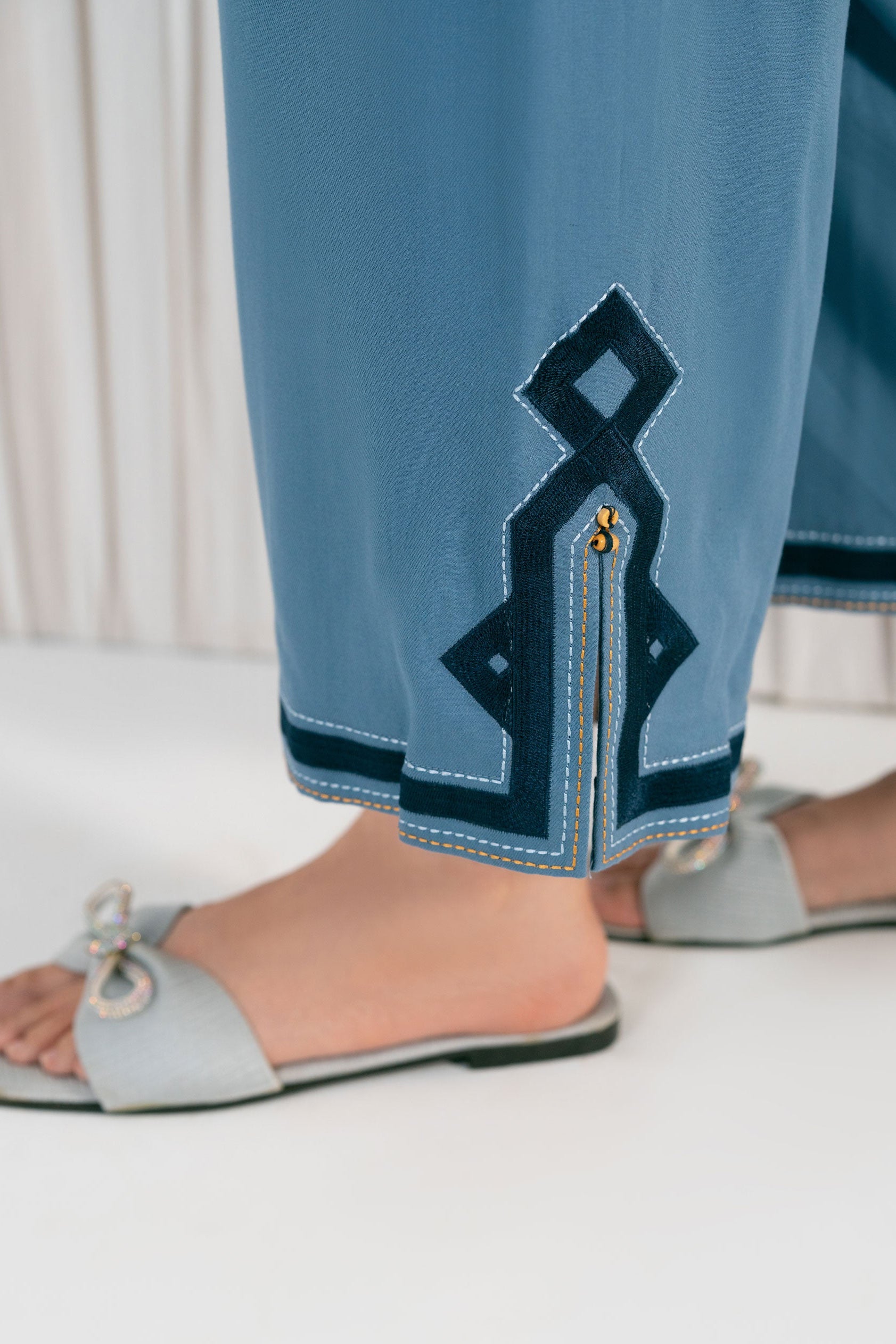 Light blue pants with decorative stitching and gray sandals on a white  background