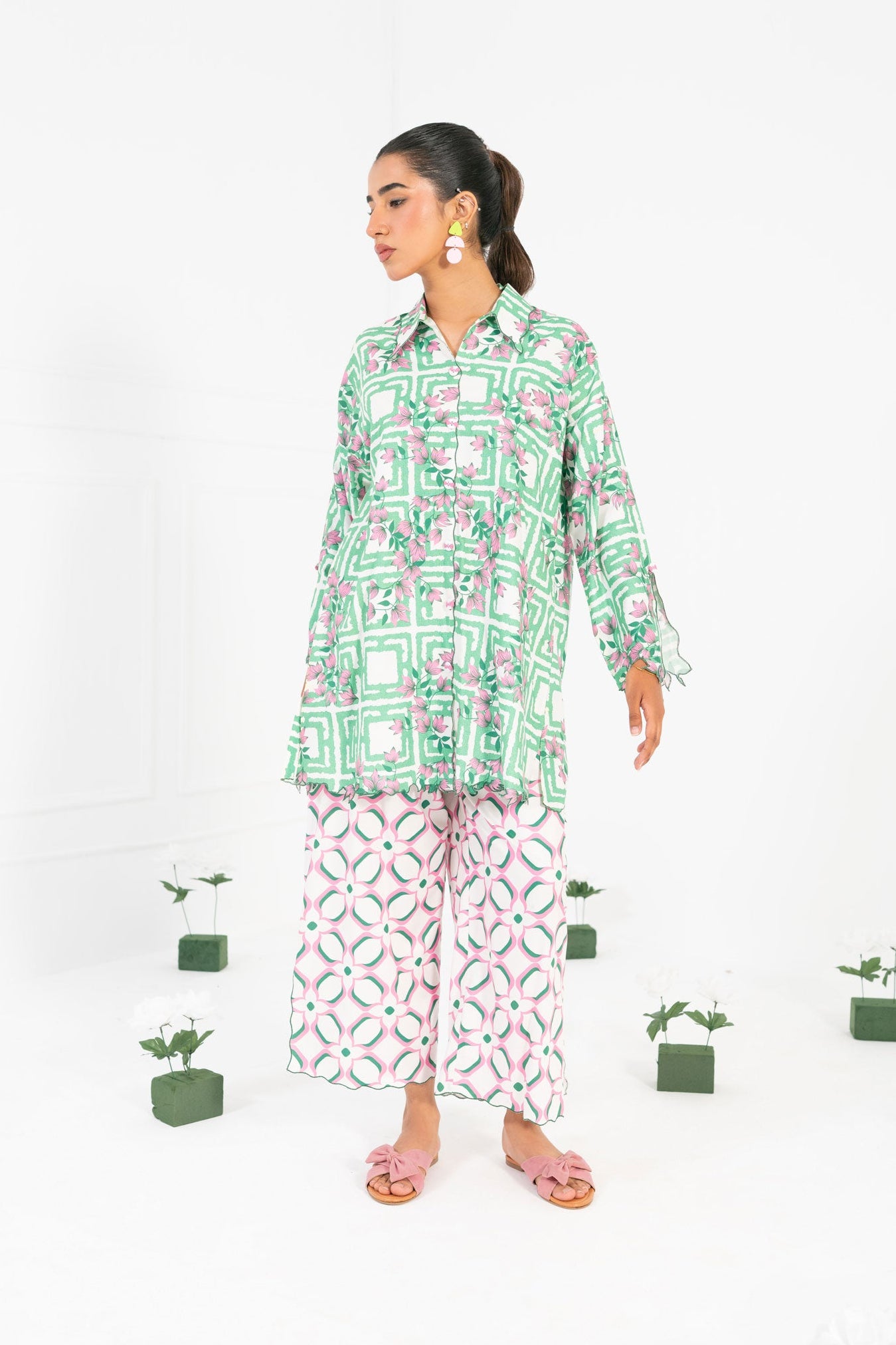 Woman wearing a green patterned shirt and white pants with black patterns, standing against a white background with small plants.