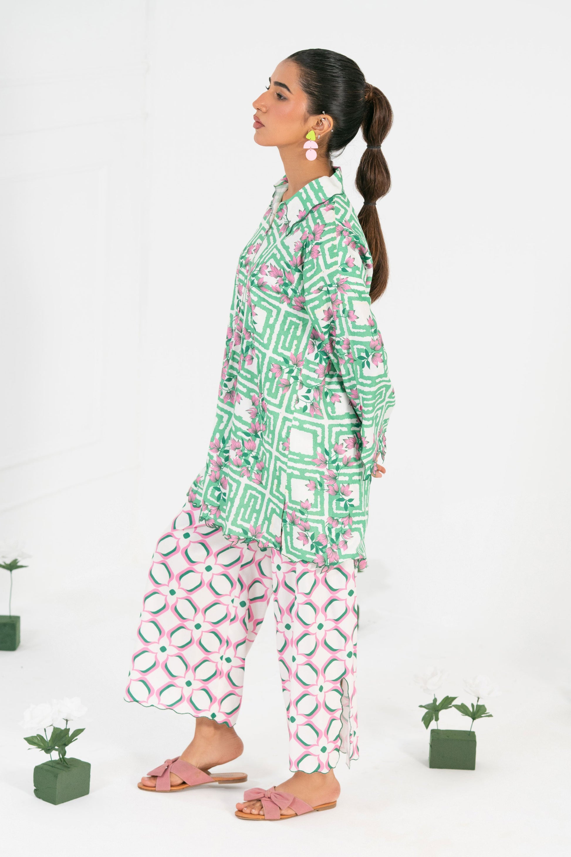 Woman wearing a green patterned top and white pants with red designs, standing against a white background with small potted plants.