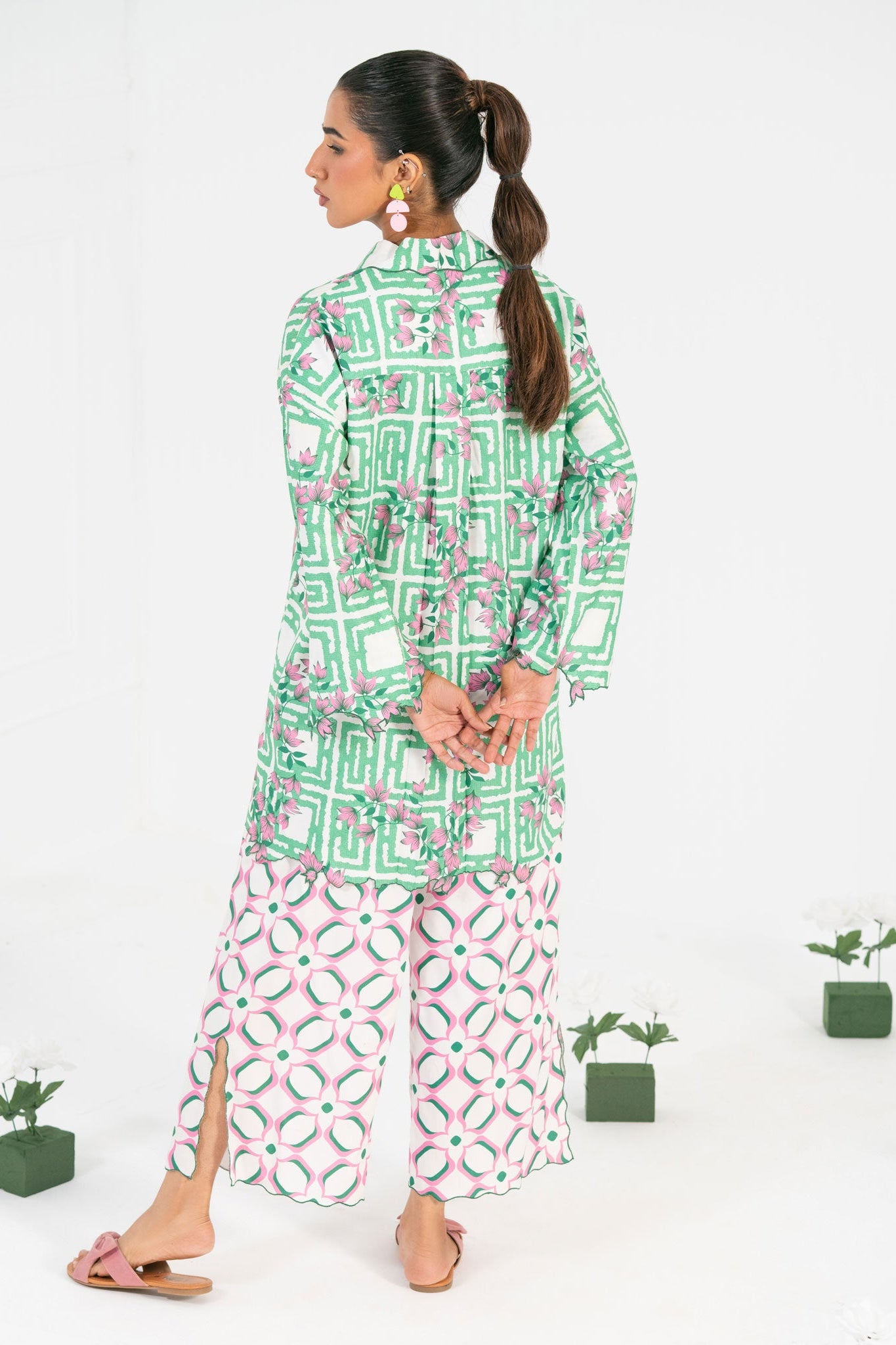 Woman wearing a green and white patterned outfit with pink pants, standing against a white background with small plants.