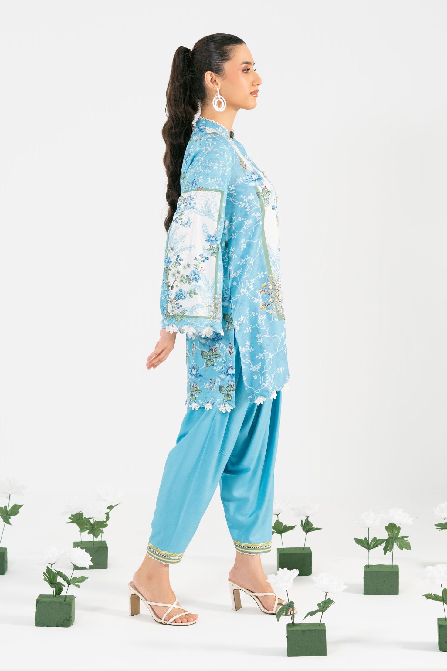 Woman in a blue floral outfit standing among potted plants on a white background