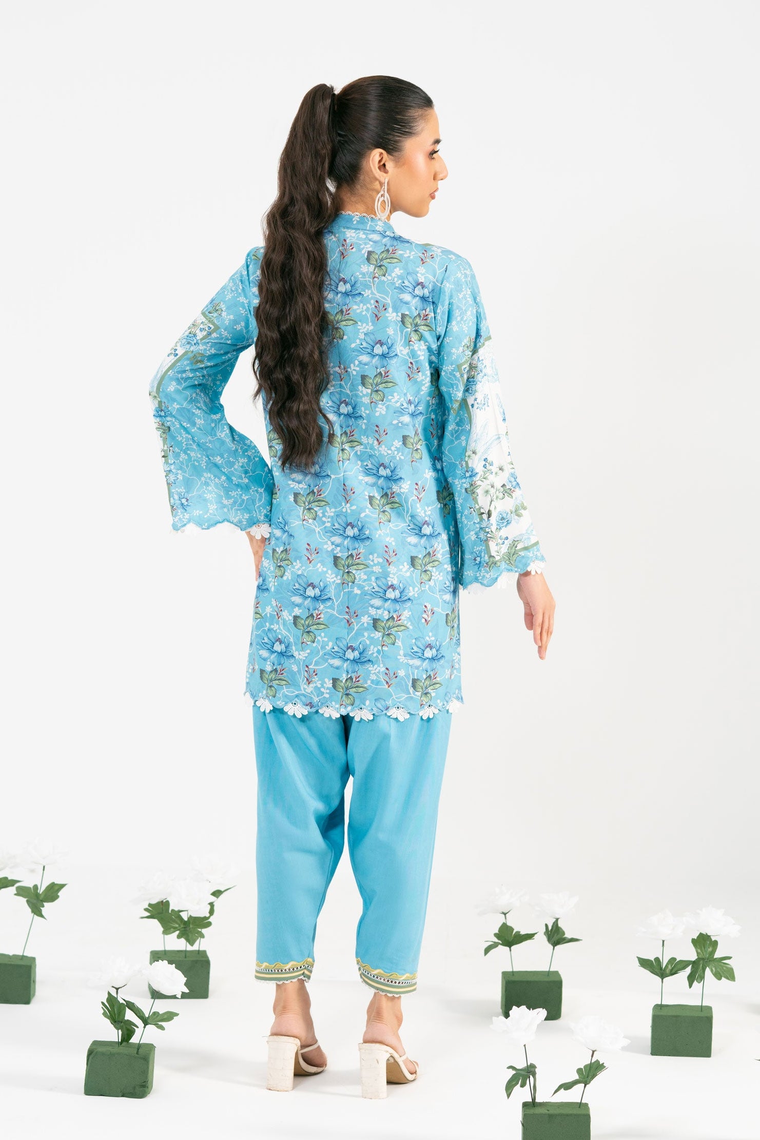 Woman in a blue floral outfit standing among potted plants on a white background