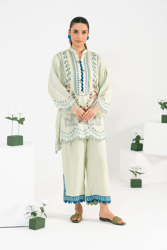 Woman wearing a traditional outfit with embroidery, standing in a minimalistic room with plants.