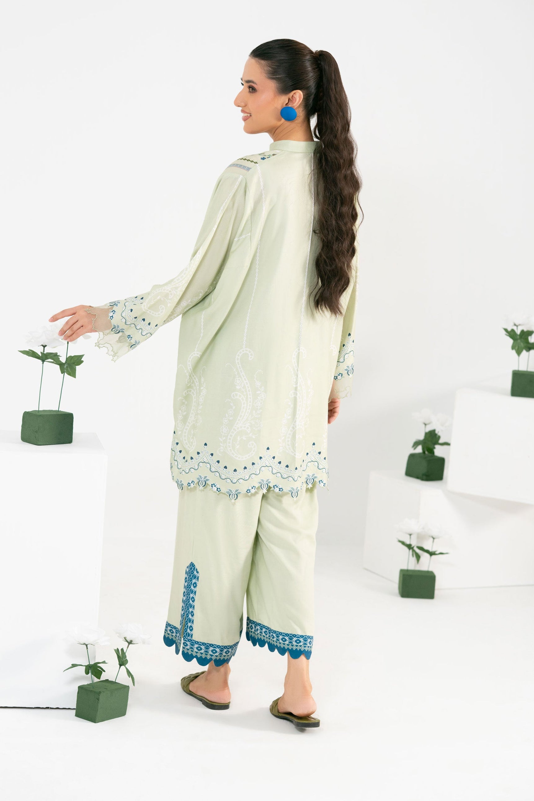 Woman in a light green traditional outfit with blue embroidery standing against a white background with potted plants.